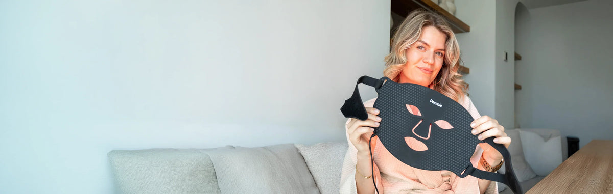 Woman holding a black Parvale LED face mask in a bright living room setting.