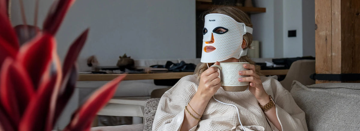 Woman wearing a white Parvale LED light therapy face mask while holding a mug in a living room.